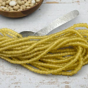 May include: A pile of yellow glass beads, with a spoon and a wooden bowl filled with white beans in the background. The beads are strung together, and the color is a soft, muted yellow. The beads are small and round, and the overall effect is delicate and pretty.