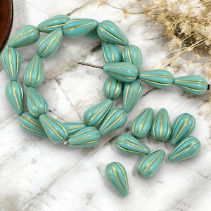 May include: Teal-colored teardrop beads with gold stripes. These beads are strung together, with some loose beads scattered around. The beads have a glossy finish and are likely for jewelry making.