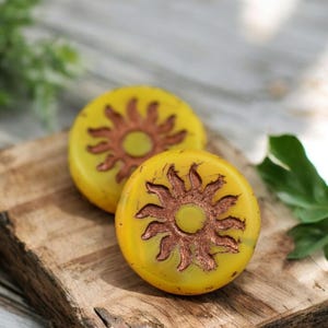 May include: Two round yellow ceramic coasters with a sun design in a copper color. The coasters are on a wooden surface.
