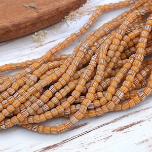 May include: Strands of orange and white striped beads. The beads are cylindrical and have a slightly irregular shape. The beads are strung together, creating a flowing, organic look. The beads are arranged on a white wooden surface.