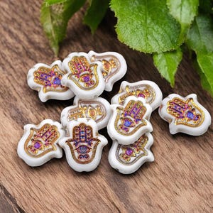 May include: A collection of white ceramic hamsa hand charms with gold and purple accents. The charms are decorated with intricate designs and feature a blue eye in the center.