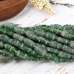 May include: Strands of green glass beads with a weathered, antique appearance. The beads are cylindrical with rounded ends and are strung together. The beads have a textured surface with white deposits, suggesting age and use. The beads are displayed on a white wooden surface.