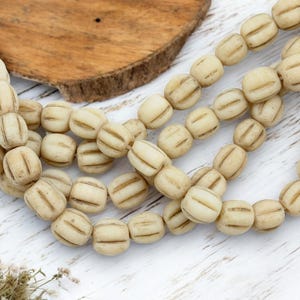 May include: Cream-colored, barrel-shaped beads with etched lines, strung together. The beads are arranged in multiple strands, suggesting a necklace or bracelet. The background includes a wooden element and a white, textured surface.