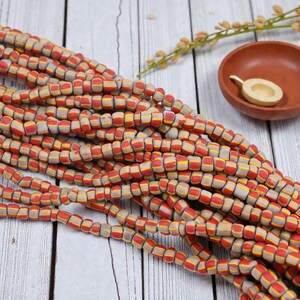 May include: Strands of colorful beads in red, yellow, and gray. The beads are small, rectangular, and strung together. A small, terracotta-colored bowl and a small wooden spoon are in the background. The beads are likely for jewelry making.