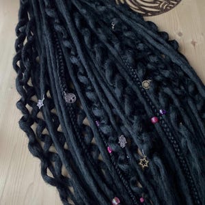 May include: Dark grey dreadlocks with decorative charms and beads. The dreadlocks feature braided and twisted sections, embellished with silver and gold charms, such as stars, hands, and tree of life pendants. Pink and purple beads are also included.