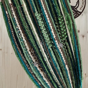 Può includere: Una collezione di dreadlock verdi, verde acqua, marroni e color crema. Alcuni dreadlock sono adornati con elementi decorativi. La disposizione è mostrata su una superficie di legno, con un logo nell'angolo in alto a destra.