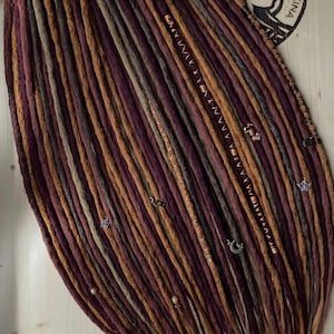 Puede incluir: Un conjunto de rastas hechas a mano en tonos morados, marrones y grises, adornadas con cuentas y abalorios decorativos. Las rastas están dispuestas en un patrón en capas, mostrando los diversos colores y texturas. Un logotipo circular con el texto "DREADKINA" es visible.