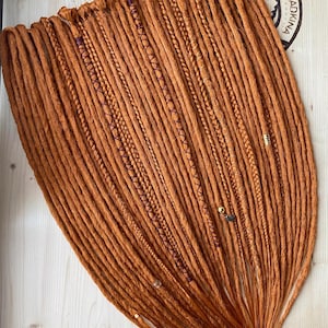 Puede incluir: Un conjunto de rastas sintéticas de color jengibre que presenta una mezcla de texturas lisas, trenzadas y envueltas. Los mechones están adornados con pequeñas cuentas metálicas y detalles de hilo oscuro, con la marca DREADKINA visible en la esquina.