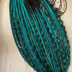 Könnte beinhalten: Ein Set aus synthetischen Dreadlocks an einem Metallring mit einem Farbverlauf von Dunkelbraun am Ansatz bis hin zu leuchtendem Türkis und Smaragdgrün an den Spitzen. Das Haar weist wellige Texturen, geflochtene Strähnen und kleine silberne Anhänger auf.