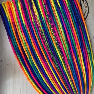 May include: A set of multi-coloured synthetic dreadlocks featuring a vibrant rainbow palette of neon orange, yellow, pink, purple, and blue strands. The collection includes smooth twisted locks and several intricate braided sections with the text DREADKINA.