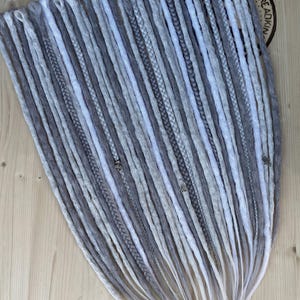May include: A collection of grey and white dreadlocks, arranged in a fan shape. The dreads vary in shades of grey, with some featuring braided sections and decorative charms. The tips fade to a lighter colour, creating a gradient effect. The background is a light wood surface.