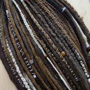 Könnte beinhalten: Nahaufnahme einer Sammlung brauner, schwarzer und weißer Dreadlocks. Einige Dreadlocks sind geflochten, andere mit weißem Faden in einem X-Muster umwickelt. Einige Dreadlocks haben Holzperlen und kleine silberne Anhänger.