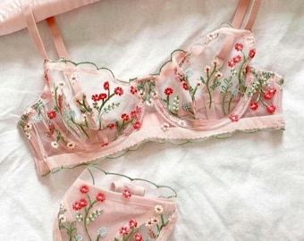 May include: A delicate pink lingerie set with floral embroidery. The bra has adjustable straps and a scalloped edge. The matching panty has a low rise and a floral design.