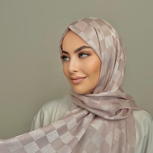 May include: A light pink checkered patterned hijab with a satin sheen. The hijab is worn by a person with light brown hair and makeup.