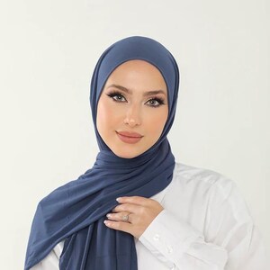 Può includere: Una donna indossa un hijab in jersey blu scuro. L'hijab è drappeggiato sulle spalle e legato al collo. Indossa una camicia bianca abbottonata.