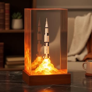 May include: A decorative lamp featuring a model rocket launching, encased in a clear acrylic block. The rocket is white and black with "USA" printed on it. The base is a dark wood, and the interior has a warm, glowing light effect.