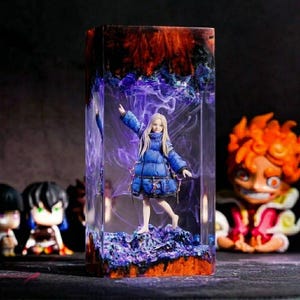 May include: A rectangular resin art piece featuring a miniature figure in a blue coat. The figure stands on a purple base, encased in clear resin with swirling purple and orange accents. Other figurines are visible in the background.