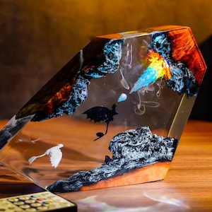 Może przedstawiać: Przezroczysta rzeźba z żywicy z wbudowaną sztuką abstrakcyjną. Grafika przedstawia czarną sylwetkę ptaka, białego ptaka i kolorowe akcenty w kolorze niebieskim, pomarańczowym i czarnym. Rzeźba jest eksponowana na drewnianej powierzchni.