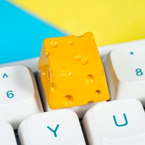 Könnte beinhalten: Ein hellgelber, käseförmiger Keycap mit Löchern liegt auf einer weißen Tastatur. Der Keycap steht im Kontrast zu den weißen Tasten und einem blau-gelben Hintergrund. Die Tastaturtasten sind mit Buchstaben und Zahlen beschriftet.