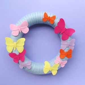 Puede incluir: Una corona de fieltro azul decorada con mariposas de fieltro de colores. Las mariposas son de varios tonos de rosa, amarillo, naranja y morado.