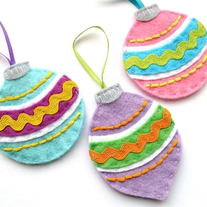 May include: Three felt Christmas ornaments in different colors. The ornaments are decorated with colorful stripes and wavy lines. The ornaments are hanging from a white background.