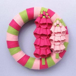 May include: A pink and green felt wreath with two pink felt flowers. The flowers are made of overlapping petals and have a textured surface.