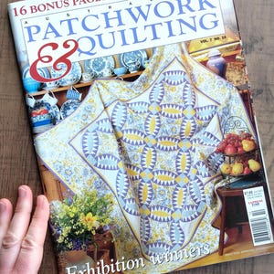 Könnte beinhalten: Ein Magazin mit dem Titel "Australian Patchwork & Quilting" mit einem Quilt auf dem Cover. Das Cover zeigt einen Quilt mit einem geometrischen Design in Gelb, Blau und Weiß. Das Magazin hat auch den Text "Exhibition winners".