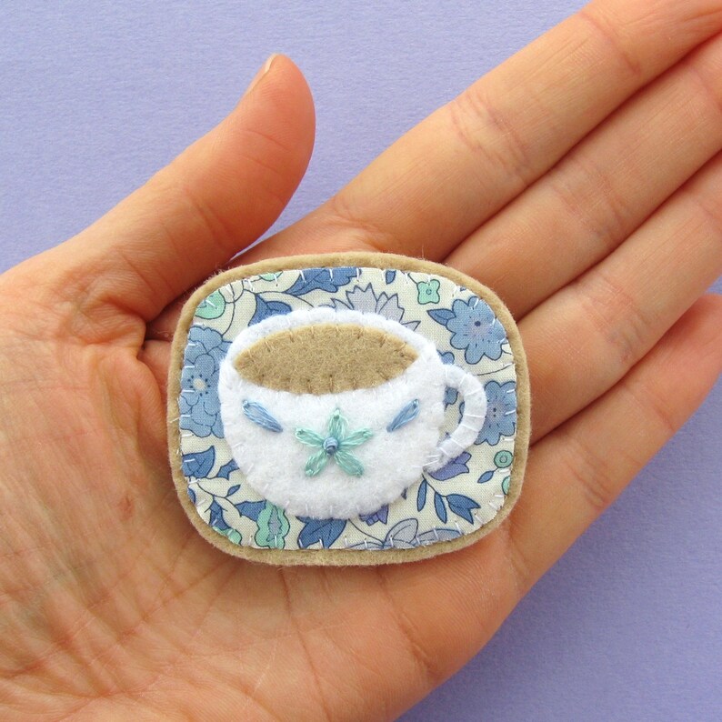 May include: A handmade felt brooch featuring a white teacup filled with brown liquid, adorned with blue embroidered details. The teacup sits on a floral patterned coaster with a light brown border. The brooch is a charming accessory.