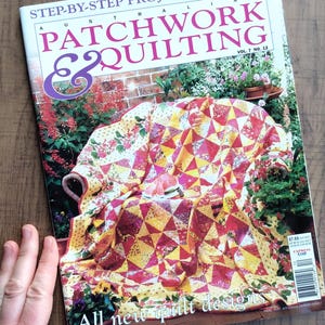 May include: A magazine titled "Australian Patchwork & Quilting" with a colourful quilt on the cover. The quilt features red, yellow, and white geometric patterns. The magazine is open on a wooden surface.