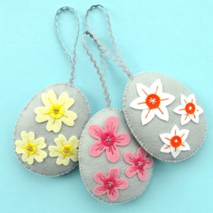 May include: Three grey felt Easter eggs with different flower designs. One egg has white flowers with orange centres, another has pink flowers with silver centres, and the third has yellow flowers with gold centres.