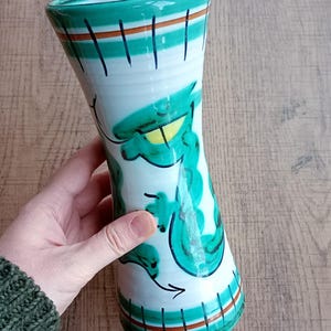 May include: A ceramic vase with a white base, decorated with green and brown stripes and a whimsical green design. The vase has a flared shape and is approximately 20 cm tall.