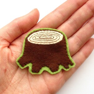 May include: A brown felt tree stump applique with a light green border. The top of the stump features a cream-colored, stitched spiral design. This handmade item is ideal for crafting and sewing projects.