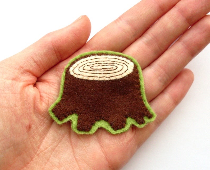 May include: A brown felt tree stump applique with a light green border. The top of the stump features a cream-colored, stitched spiral design. This handmade item is ideal for crafting and sewing projects.