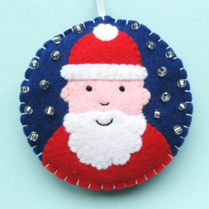 May include: A felt Christmas ornament featuring a Santa Claus face with a white beard and red hat. The ornament is decorated with white beads on a blue background.