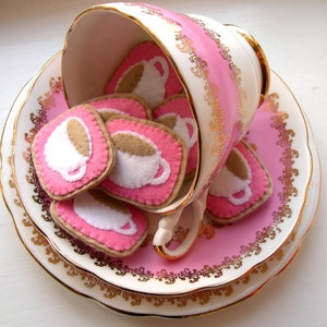 May include: A vintage teacup and saucer set in pink and white, adorned with gold trim. The teacup is overflowing with felt coasters, each featuring a coffee cup design in white and brown on a pink background.
