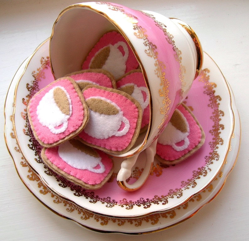 May include: A vintage teacup and saucer set in pink and white, adorned with gold trim. The teacup is overflowing with felt coasters, each featuring a coffee cup design in white and brown on a pink background.