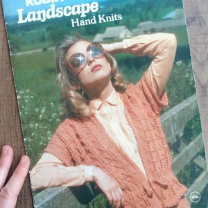 May include: A vintage book cover for "Robin Landscape Hand Knits". It shows a woman in sunglasses wearing a peach-coloured knitted vest. The background features a green field and a blue sky.