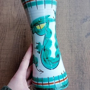 May include: A white ceramic vase with a turquoise dragon design. The vase has a flared shape and is decorated with black and brown stripes. The dragon has yellow eyes and is outlined in black. The vase is held by a person.