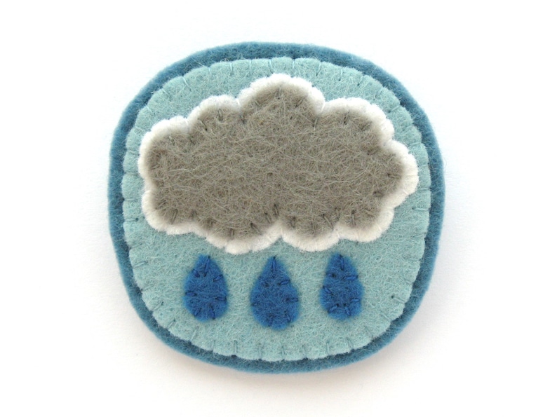 May include: A felt brooch featuring a whimsical weather design. The brooch has a light blue background with a gray cloud and three blue raindrops. The cloud has white accents, and the brooch is round.