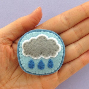 May include: A handmade felt pin featuring a gray cloud with white trim and three blue raindrops. The pin is round, with a light blue background and a darker blue border. The pin is approximately 2 inches in diameter.