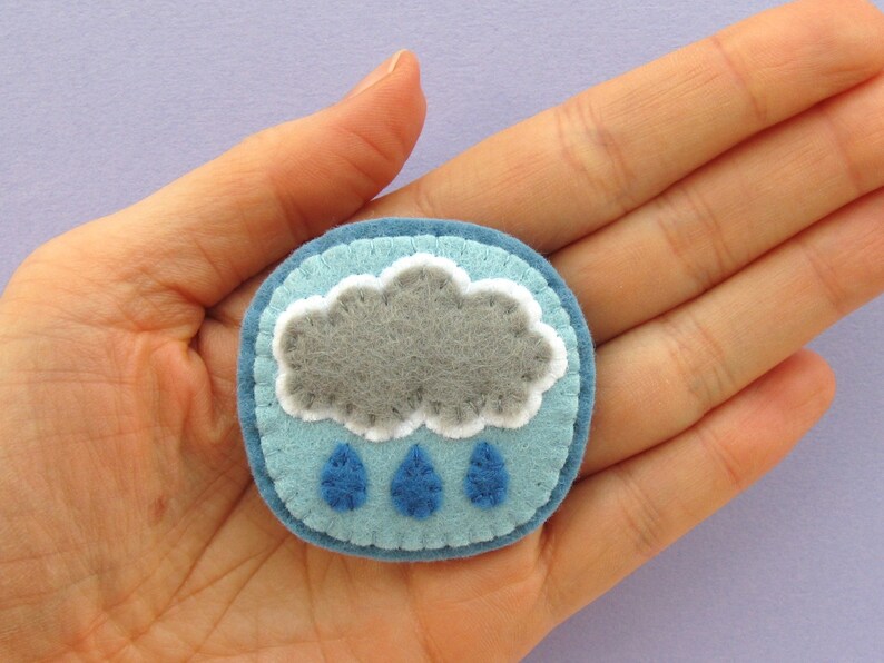 May include: A handmade felt pin featuring a gray cloud with white trim and three blue raindrops. The pin is round, with a light blue background and a darker blue border. The pin is approximately 2 inches in diameter.