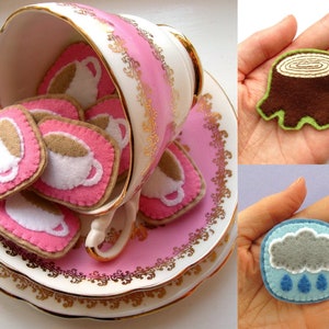 May include: A pink teacup and saucer overflowing with felt coffee cup-shaped cookies. The cookies have a pink border, a tan cookie base, and a white coffee cup design. Two felt pins, one a tree stump and the other a rain cloud, are also shown.
