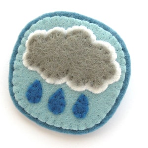 May include: A round, felt pin featuring a gray cloud with white trim and three blue raindrops. The cloud and raindrops are on a light blue background, with a darker blue border. The pin is handmade.