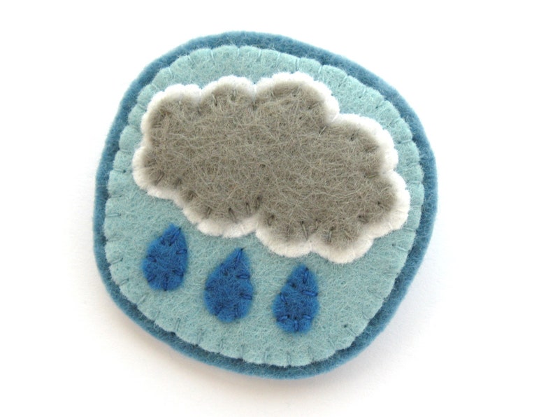May include: A round, felt pin featuring a gray cloud with white trim and three blue raindrops. The cloud and raindrops are on a light blue background, with a darker blue border. The pin is handmade.