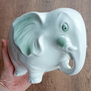May include: A white ceramic elephant planter with green detailing on the ears and face. The elephant has a trunk, small legs, and a rounded body. The planter is designed for small plants or flowers.