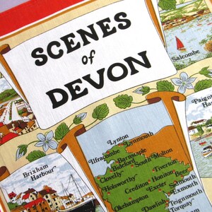 May include: A decorative tea towel featuring the text SCENES of DEVON in bold black letters. The fabric displays a map of Devon with various town names, alongside illustrated coastal scenes of Brixham Harbour, Salcombe, and Paignton Harbour, accented with green floral motifs and a red border.