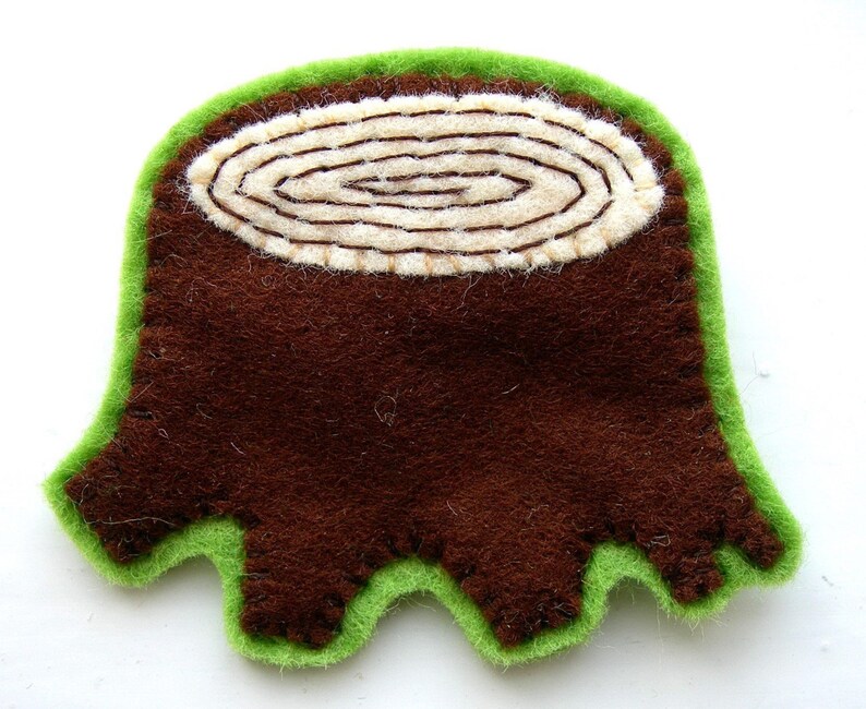 May include: A handmade felt tree stump applique. The tree stump is brown with a cream-colored center showing the growth rings. The applique is outlined with green felt, giving it a three-dimensional appearance. This item is suitable for crafting and sewing projects.