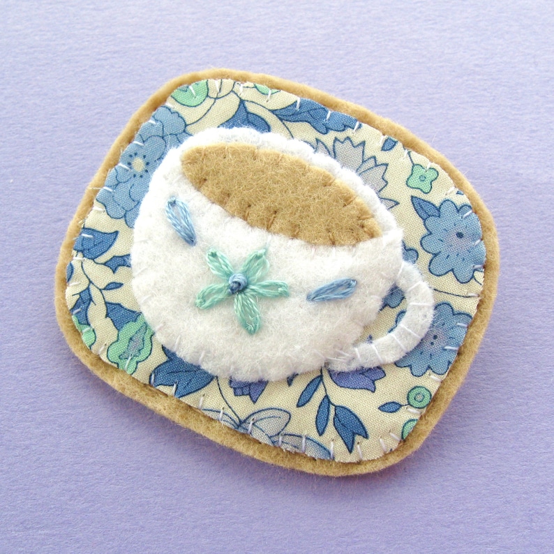 May include: A handmade felt brooch featuring a white teacup with a tan-colored tea filling, adorned with a light blue embroidered flower and accents. The teacup sits on a floral patterned coaster with a tan border.