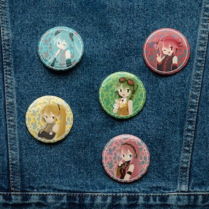 May include: Five circular pin-back buttons featuring anime-style characters. The buttons display characters with teal, red, green, yellow, and pink hair, each set against a unique patterned background in coordinating colors, arranged on blue denim fabric.