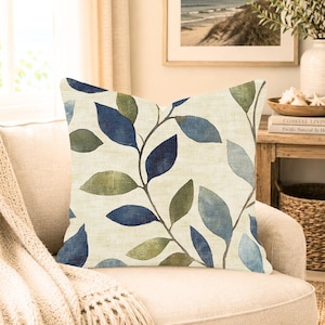 May include: A square throw pillow featuring a cream-colored background with a repeating botanical pattern of leaves in shades of navy blue, sage green, and light blue. The fabric has a visible woven texture, and the leaves are arranged on thin stems.